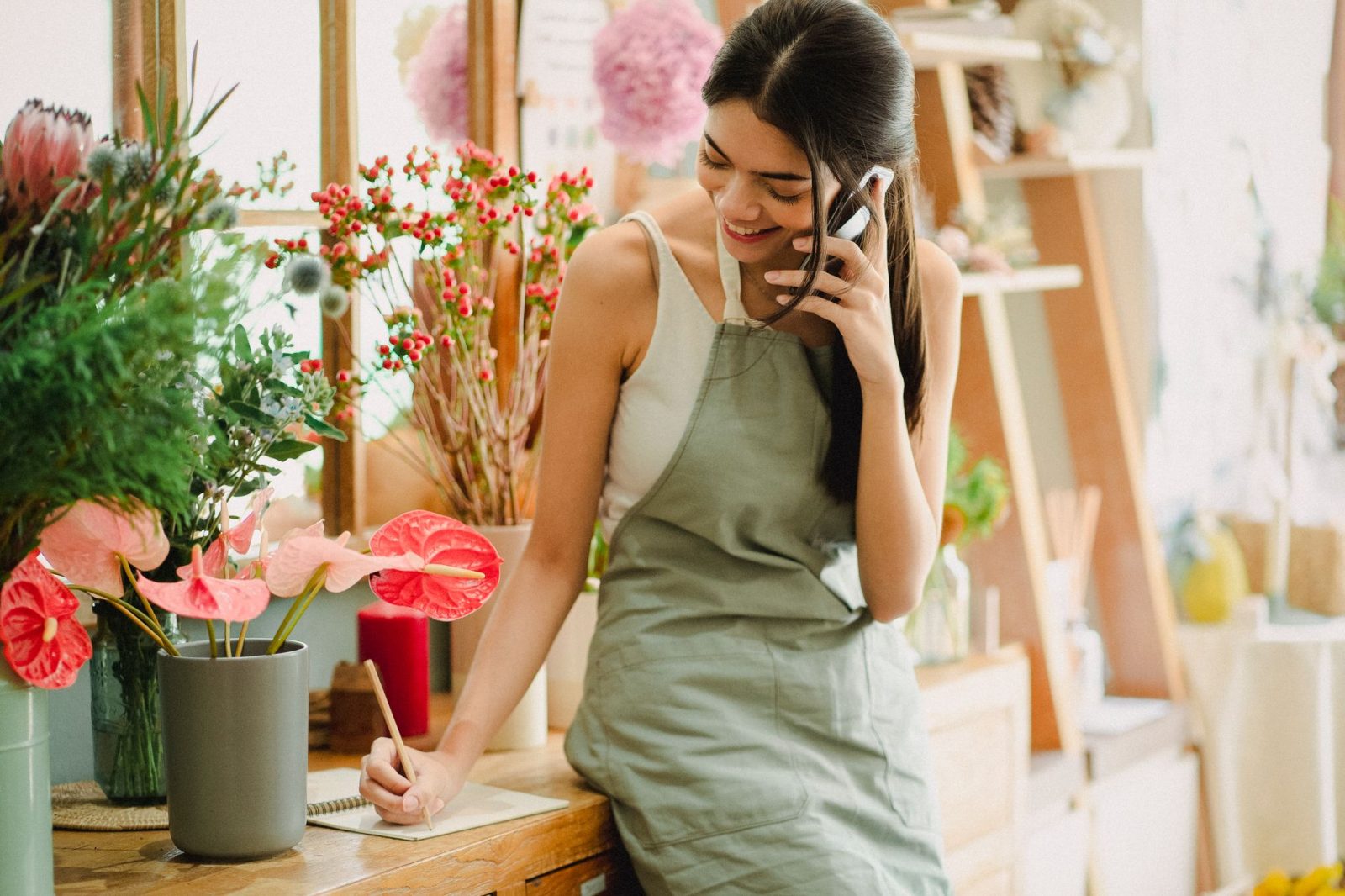 Florist in an apron taking an order on her phone in her shop while writing it down on a notepad